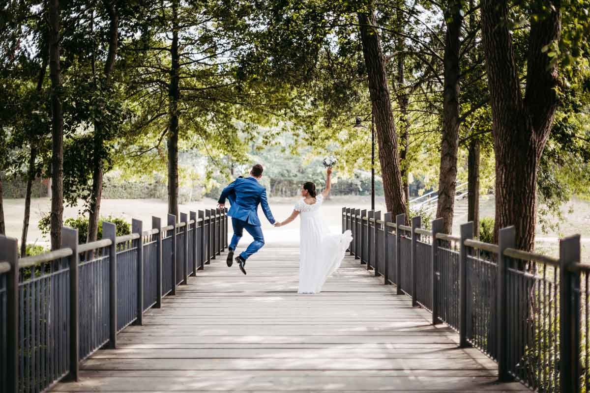 Brautpaar gehen Hand in Hand über die Brücke. Bräutigam springt nach oben.